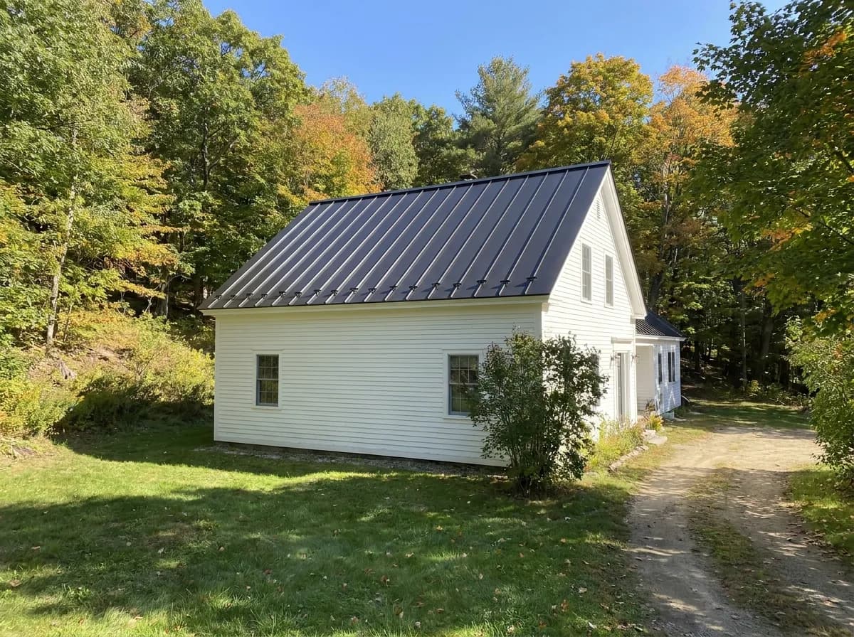 White New England farmhouse on a wooded hillside with a new dark standing-seam metal roof and snow guards along the eave
